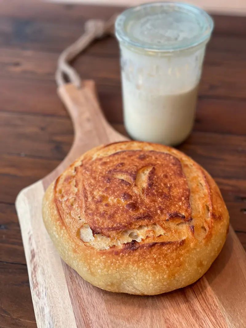 Pain rond sur une planche, avec un pot de levain à l’arrière-plan